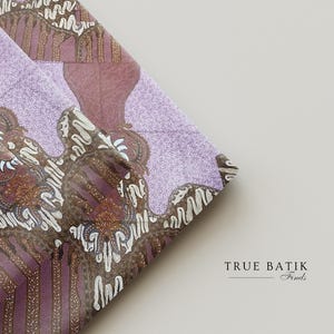 Op de afbeelding: Close-up van gevouwen stof met een batikpatroon. De stof heeft een mix van paarse, bruine en witte kleuren. Het ontwerp bevat bloemen en geometrische elementen. De tekst "TRUE BATIK Finds" staat rechtsonder.