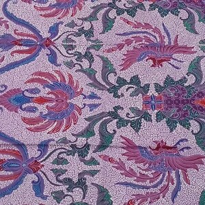 Op de afbeelding: Een gedetailleerd textiel met een herhaald patroon van gestileerde bloemen- en vogelmotieven in tinten paars, blauw, groen en rood op een lichtpaarse achtergrond. De stof heeft een getextureerd uiterlijk, wat wijst op een geweven of bedrukt ontwerp.