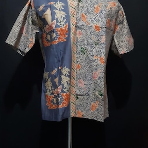 Peut inclure: Une chemise à manches courtes avec un motif batik unique. La chemise présente un design divisé avec un panneau bleu orné de motifs floraux et de palmiers, et un panneau à motifs avec des accents orange et verts.