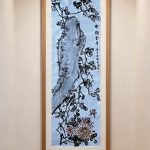 May include: A vertical framed ink wash painting featuring a large grey rock formation, black ink branches, and pink and yellow chrysanthemum flowers on a light blue background. The artwork includes black Chinese calligraphy and measures 34x136 cm.