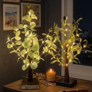 Puede incluir: Dos árboles artificiales decorativos con luces LED blancas cálidas. Uno tiene hojas verdes redondas, el otro tiene hojas parecidas a las de la oliva y pequeños detalles negros. Ambos tienen troncos marrones y se asientan sobre bases cuadradas.