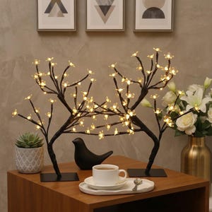 May include: Two black decorative LED cherry blossom trees with warm white lights. The trees are on a wooden table with a black bird figurine, a cup of coffee, and a small succulent plant in a patterned pot.