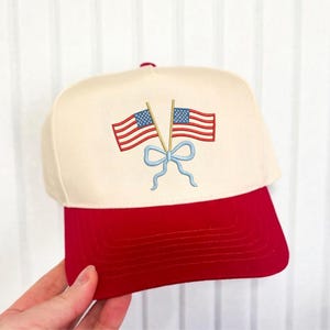 Può includere: Cappellino da baseball con corona color crema e visiera rossa. Il cappellino presenta un disegno ricamato di due bandiere americane incrociate con un fiocco azzurro. Il disegno è centrato sulla parte anteriore del cappellino.