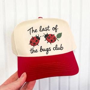 May include: A baseball cap with a cream-colored crown and a red brim. The cap features the text "The last of the bugs club" with two ladybug illustrations. The ladybugs are red with black spots and are next to a green leaf.