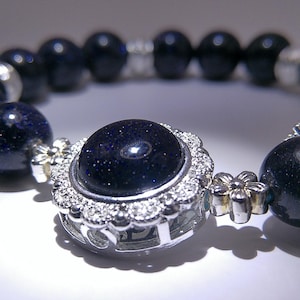 May include: A close-up of a dark blue beaded bracelet. The bracelet features large, round, dark blue beads with a sparkling effect. A central, larger bead is surrounded by a silver-colored, flower-shaped setting with clear stones.