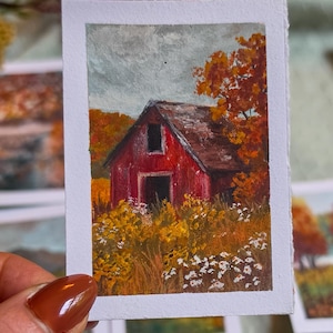 Könnte beinhalten: Ein kleines rechteckiges Gemälde auf weißem Papier, das eine rote Scheune umgeben von herbstlichem Laub zeigt. Die Szene umfasst einen grauen Himmel, orangefarbene und gelbe Bäume sowie ein Feld mit goldenen Wildblumen und kleinen weißen Blüten.