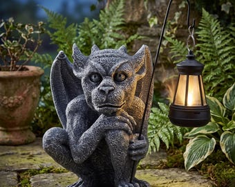 May include: A gray stone-textured gargoyle statue in a crouching position with folded wings and pointed ears. The figure holds a thin black metal shepherd's hook that suspends a small black lantern with a glowing warm light inside the glass panes.