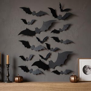 May include: A collection of black paper bat cutouts arranged in a scattered pattern on a dark gray wall. The bats vary in size and wing shape, creating a layered effect. Below the bats are two metallic pumpkins and a framed moon print.