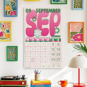 May include: A wall calendar for September with large pink lettering and floral accents. The calendar is surrounded by framed floral art in various colors. A record player, books, and a lamp are visible below.