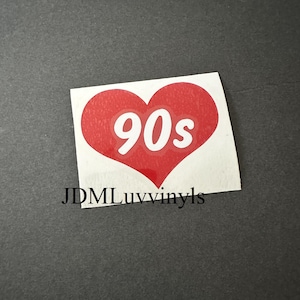 May include: A sticker featuring a red heart with the white text "90s" in the center. The sticker also includes the text "JDMLuvvinyls" at the bottom. The sticker is on a gray background.