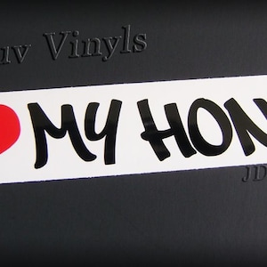 May include: White rectangular sticker with the text "I ❤️ MY HONDA." The heart is red, and the text is in black, stylized lettering. The sticker is on a black background. The words "JDMluv Vinyls" are at the top and bottom.