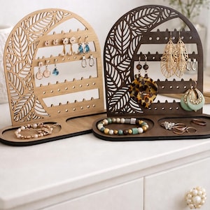 May include: Two wooden earring and jewelry organizers. One is light brown, the other is dark brown. Both have a leaf design cut out. The light brown organizer holds earrings and a bracelet. The dark brown organizer holds earrings, a necklace, bracelets, and rings.