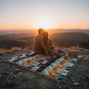 May include: A colorful woven blanket with a geometric pattern is spread on a rocky hilltop. A couple is seated on the blanket, embracing and watching the sunset. A picnic basket and mugs are nearby, creating a serene outdoor scene.