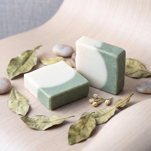 May include: Two rectangular bars of handmade soap, one with a white and green swirl design, the other with a white and green half-moon design. The soaps are surrounded by dried leaves and small stones on a light wood surface.