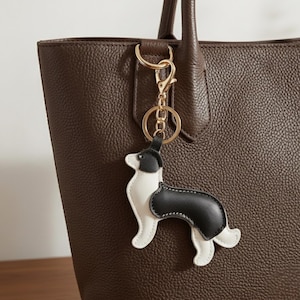May include: A black and white dog-shaped keychain with a gold-colored clasp hangs from a brown leather tote bag. The keychain is a stylized representation of a dog, with white and black sections. The bag has a textured surface.