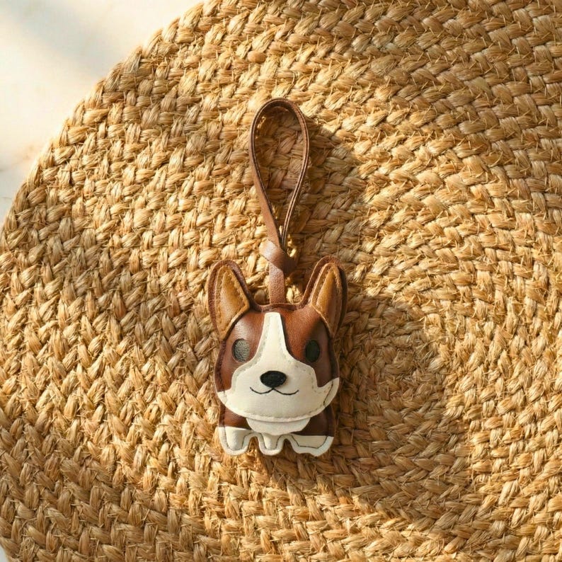 May include: A brown, white, and tan leather corgi keychain with a brown leather loop. The corgi keychain is lying on a woven, tan, circular mat.