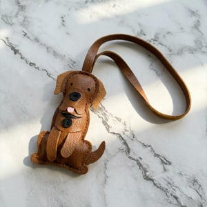 May include: A brown leather dog-shaped keychain with a loop. The dog has a black nose, button eyes, and a pink tongue. The keychain is made of textured leather and is sitting on a marble surface.
