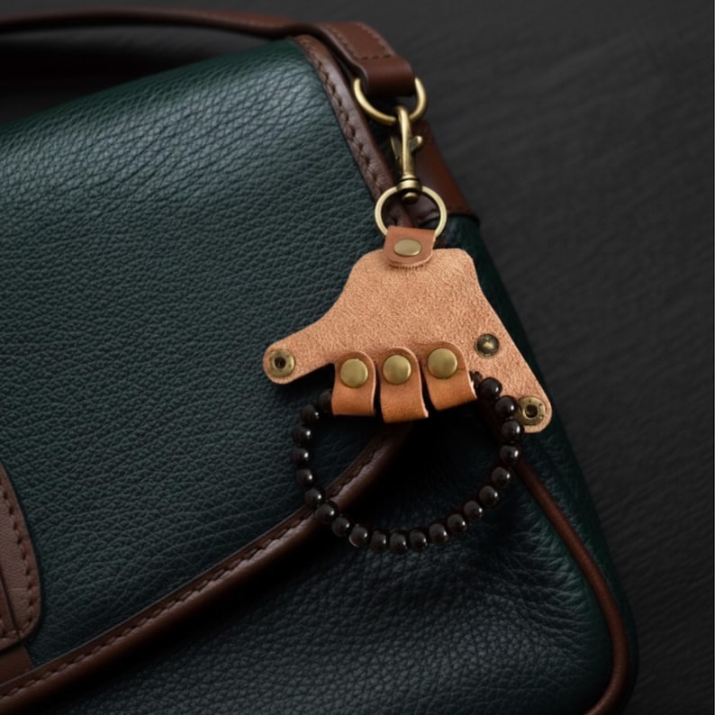 May include: A leather bag charm with a light brown leather hand design, brass rivets, and a black beaded bracelet. The charm is attached to a green leather bag with brown trim and a brass clasp.