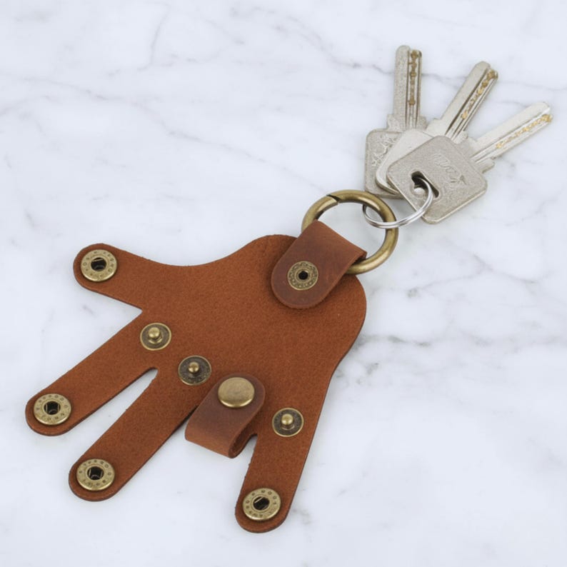 May include: A brown leather keychain in the shape of a hand, with brass rivets and a brass ring holding several keys. The keychain is made of brown leather and has a unique design.