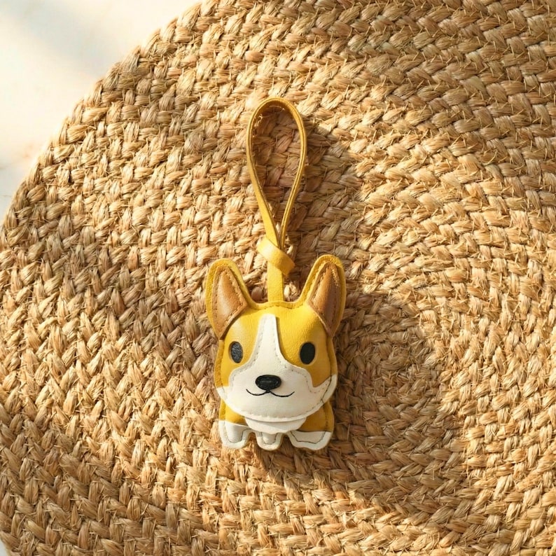 May include: A yellow and white corgi-shaped keychain with a matching yellow loop. The keychain has black eyes and a black nose. The background is a woven, tan mat.