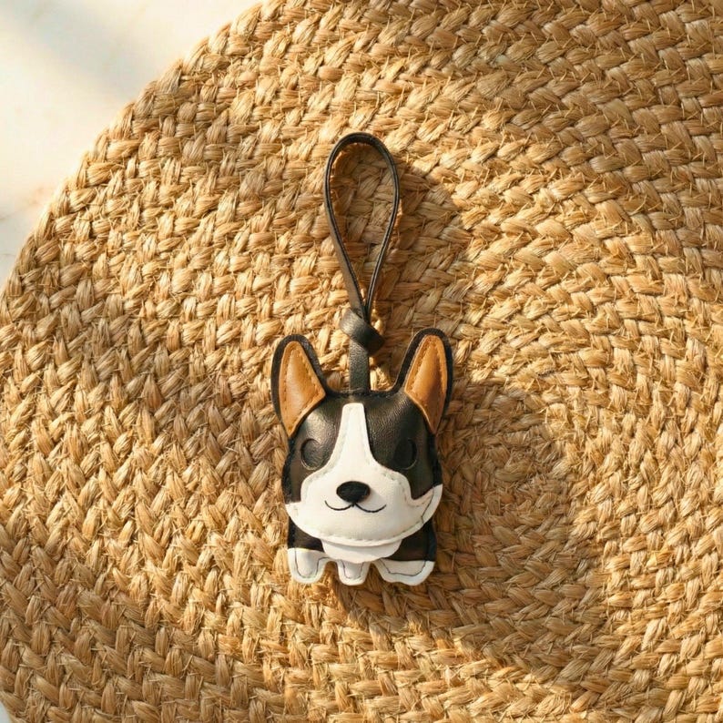 May include: A black, white, and tan corgi-shaped keychain with a black leather loop. The keychain is made of leather and has a smiling face. The background is a woven, tan mat.