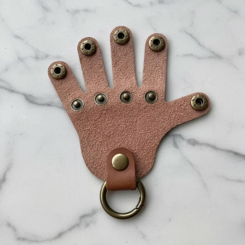 May include: A pink leather keychain shaped like a hand, featuring five finger-shaped cutouts. The keychain has metal snap closures on each finger and a metal ring attached to a leather loop at the base. The background is a white marble.