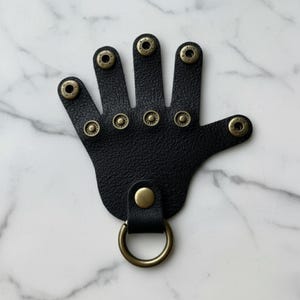 May include: Black leather hand-shaped keychain with brass-colored rivets and a brass-colored ring. The keychain has a textured surface and a loop for attaching keys or other items. The design is a unique and stylish accessory.