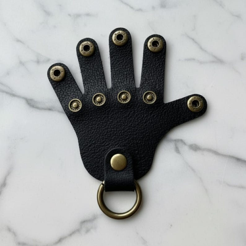 May include: Black leather hand-shaped keychain with brass-colored rivets and a brass-colored ring. The keychain has a textured surface and a loop for attaching keys or other items. The design is a unique and stylish accessory.