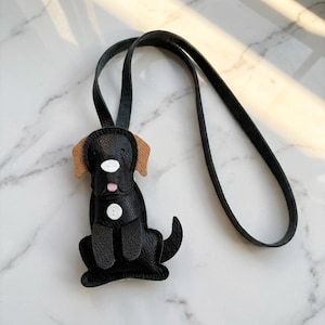 May include: A black leather dog-shaped pendant with tan ears and a pink tongue. The pendant has two white button-like accents and a black leather strap for wearing. The dog is sitting, with its tail curled.