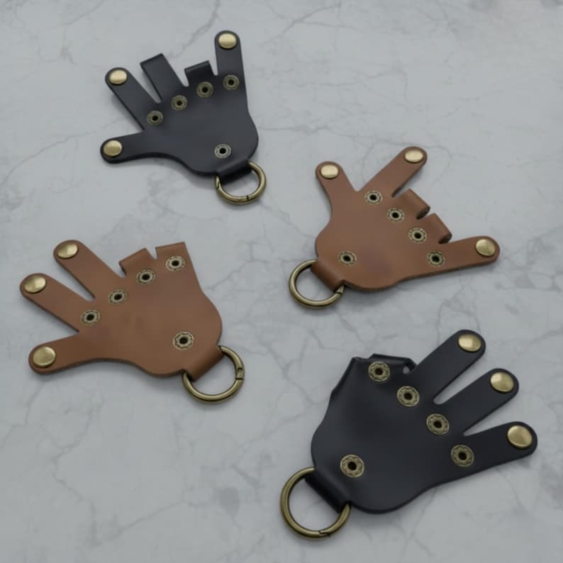 May include: Four leather hand-shaped keychains in black and brown. Each keychain has brass-colored rivets and a metal ring for attaching keys. The hands are in different gestures, including the "I love you" sign.
