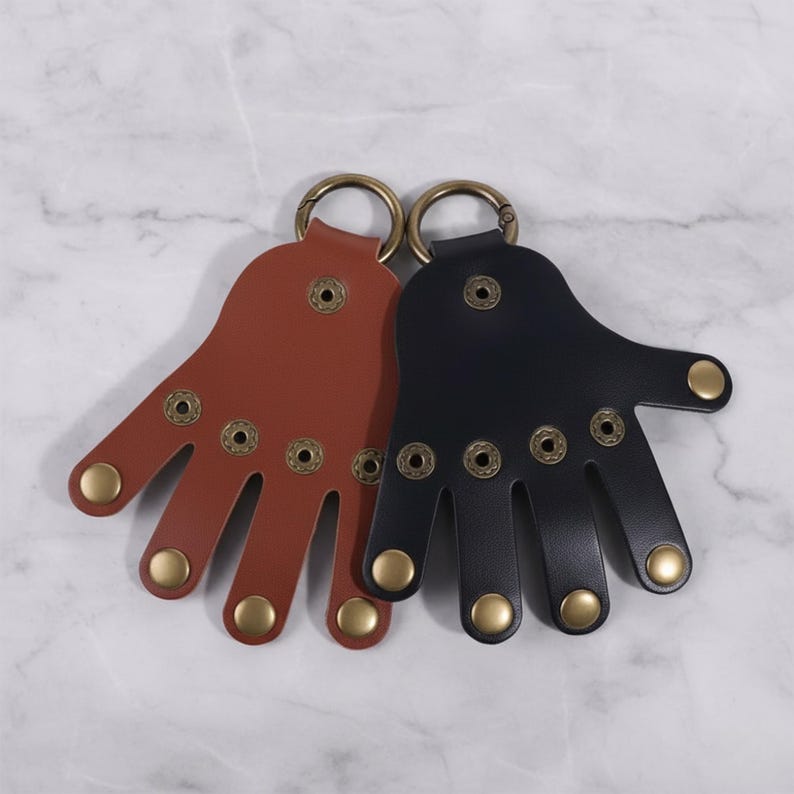 May include: Two leather hand-shaped keychains. One is brown, the other is black. Each has brass-colored rivets and a matching ring for keys. The keychains are designed to resemble a hand with five fingers.