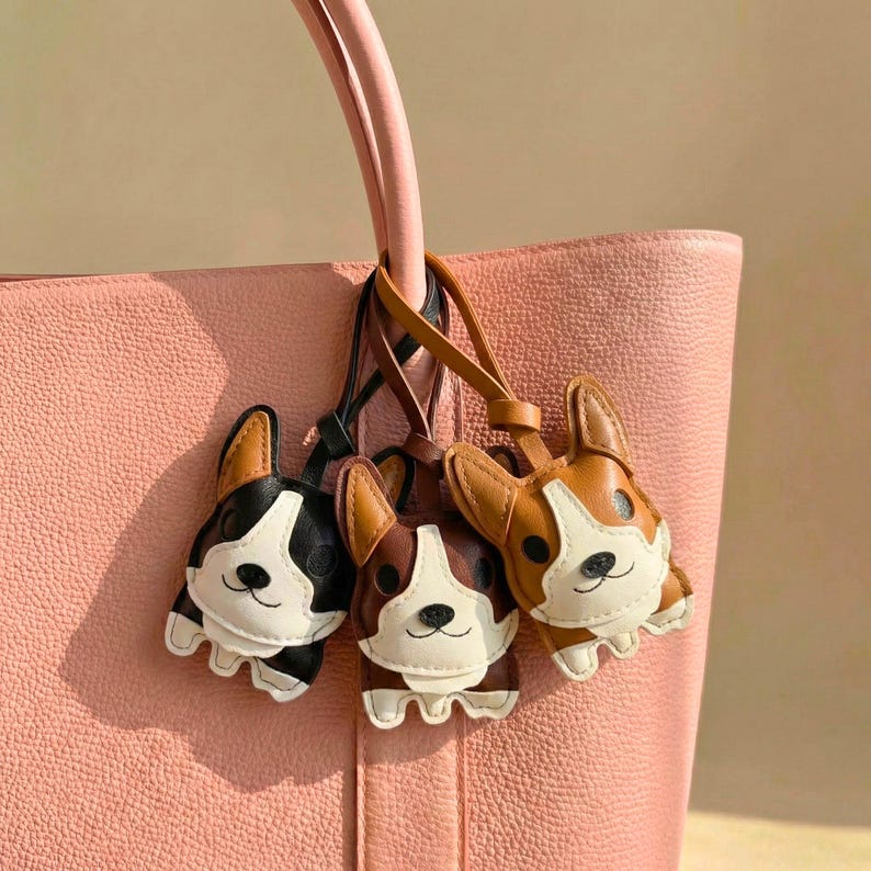 May include: Three leather dog-shaped keychains in black and white, brown and white, and brown and tan, hanging from a pink tote bag handle. The keychains have white faces and are detailed with stitched features.