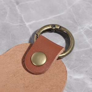 May include: Close-up of a leather keychain with a brass-colored clasp and button. The keychain features a tan leather base and a brown leather strap. The clasp is circular and attached to the strap with a brass-colored button.