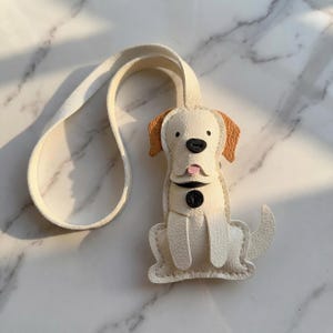 May include: A cream-colored leather dog-shaped keychain with tan ears and a black nose. The dog has a pink tongue and a black button collar. A matching cream-colored strap extends from the top of the keychain.