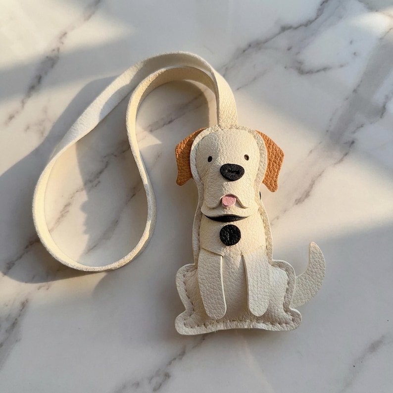 May include: A cream-colored leather dog-shaped keychain with tan ears and a black nose. The dog has a pink tongue and a black button collar. A matching cream-colored strap extends from the top of the keychain.