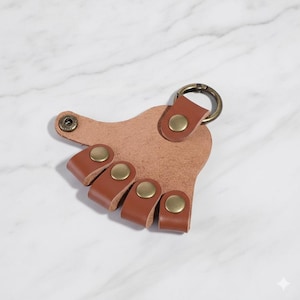May include: A tan leather keychain with four brown leather loops, each secured with a brass button. The keychain has a brass ring for attaching keys. The design is simple and functional.