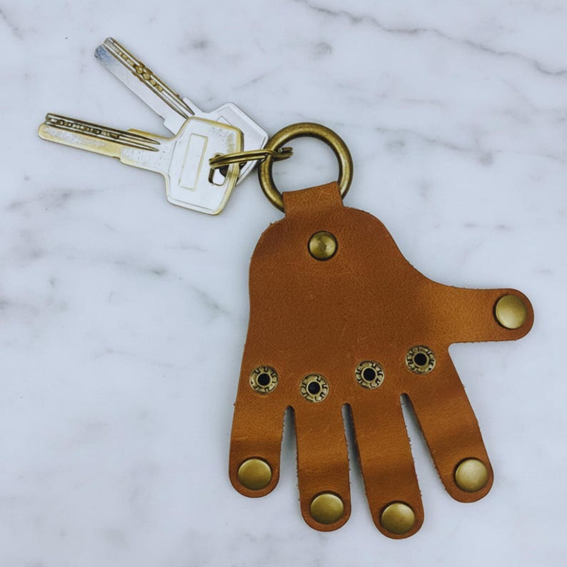 May include: A brown leather hand-shaped keychain with a brass ring and rivets. Two keys are attached to the ring. The keychain is on a white and gray marble surface.