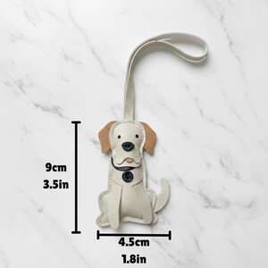 May include: A cream-colored dog-shaped keychain with tan ears and a black button nose. The keychain is 3.5 inches tall and 1.8 inches wide. It has a loop at the top for attaching to keys or a bag.