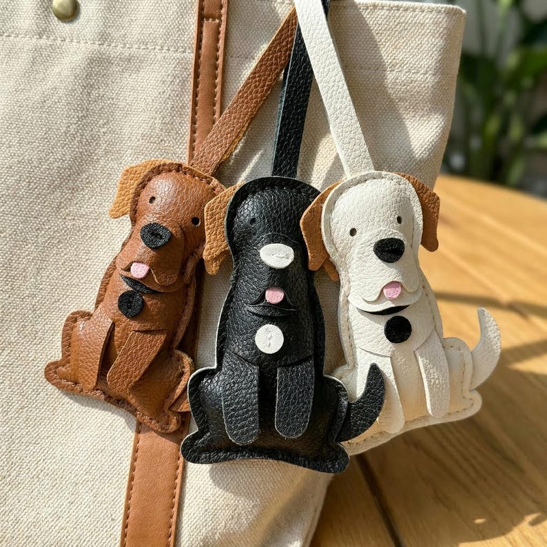May include: Three leather dog-shaped keychains in brown, black, and white. Each keychain features a dog with floppy ears, a black nose, and a pink tongue. The keychains are attached to a beige tote bag with brown leather straps.