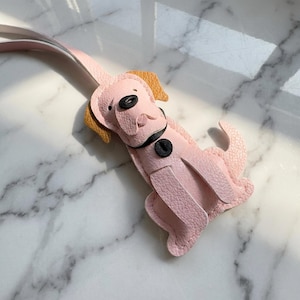 May include: A pink leather dog-shaped keychain with a light pink strap. The dog has black eyes and a black collar with a black button. The dog's ears are tan.