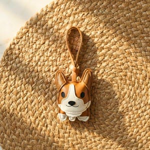 May include: A brown and white leather corgi keychain with a loop for hanging. The keychain is in the shape of a corgi dog with a smiling face. The keychain is on a woven, tan surface.