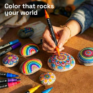 May include: The image shows painted rocks with colorful designs, including rainbows and floral patterns. An orange and black dual-tip marker is used to add details to a rock. The text "Color that rocks your world" is at the top. Other markers are visible.