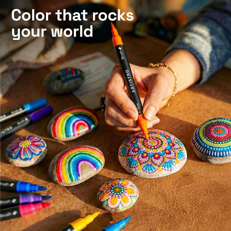 May include: The image shows painted rocks with colorful designs, including rainbows and floral patterns. An orange and black dual-tip marker is used to add details to a rock. The text "Color that rocks your world" is at the top. Other markers are visible.