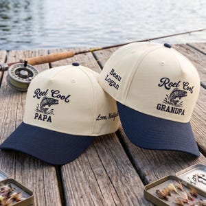 May include: Two cream-colored baseball caps with navy blue brims. The left cap features the text "Reel Cool PAPA" with a fish graphic. The right cap features "Reel Cool GRANDPA" with a fish graphic and the side text "Beau Logan" and "Love, Kailyn".