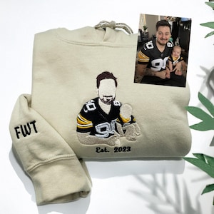 May include: A beige hooded sweatshirt featuring custom embroidery of two figures in black and yellow sports jerseys. The sleeve is embroidered with the letters FWT, and the text Est. 2023 is stitched below the central figures. A reference photo is included.