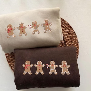 Może przedstawiać: Dwie złożone bluzy, jedna beżowa i jedna brązowa, każda haftowana czterema postaciami ludzików z piernika. Piernikowe ludziki mają czerwone szaliki, muszki i laski cukrowe. Bluzy leżą na okrągłej, tkanej macie.