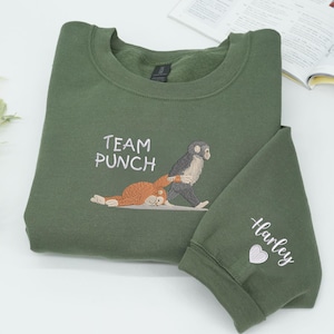 May include: Olive green sweatshirt with embroidered design. The design features the text "TEAM PUNCH" and an illustration of two monkeys. The sleeve has the name "Harley" and a heart embroidered on it.
