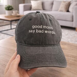 May include: A charcoal gray baseball cap with the embroidered text "good moms say bad words." The hat has a curved brim and a fabric strap closure. The hat is made of a soft, worn material.