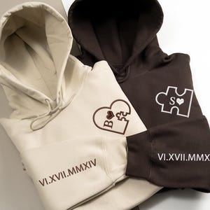 May include: Two hooded sweatshirts, one cream and one brown, are displayed. The cream sweatshirt has a heart-shaped puzzle piece design with the letters "B" and "S" embroidered in brown. The brown sweatshirt has a puzzle piece design with the letters "S" and a heart. Both have dates.