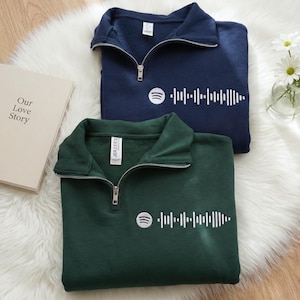 Puede incluir: Dos sudaderas con cremallera de cuarto de círculo, una azul marino y otra verde oscuro, con el logotipo blanco de Spotify y un gráfico de forma de onda de audio correspondiente. Las sudaderas están dobladas sobre una superficie de piel sintética blanca.